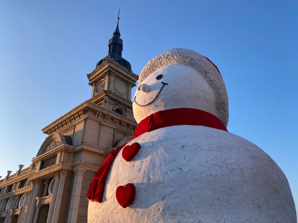 A giant snowman stands beside a historic clock tower in Harbin, China, under clear winter skies.